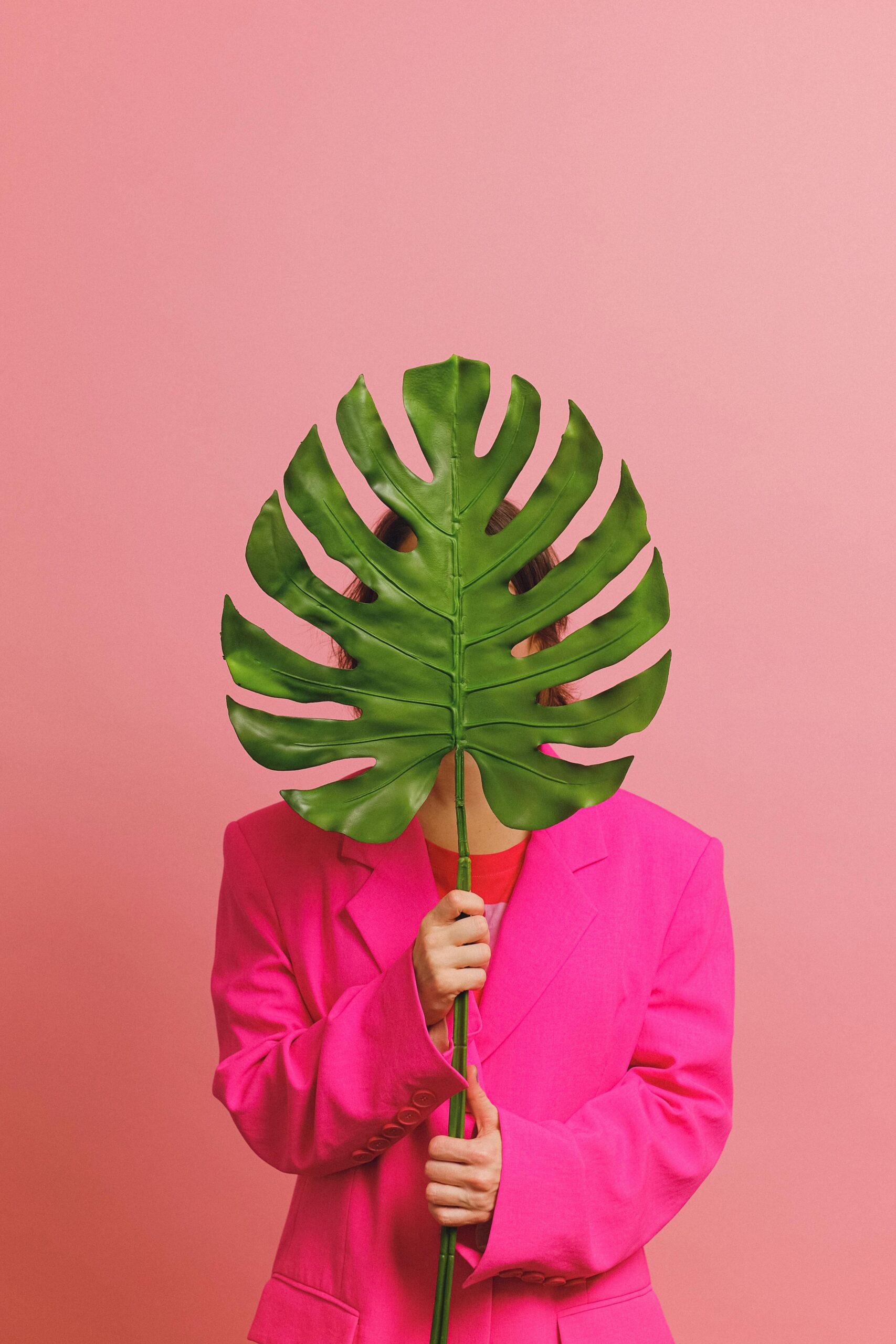 A person in a pink coat holding a large Monstera leaf against a solid pink background.